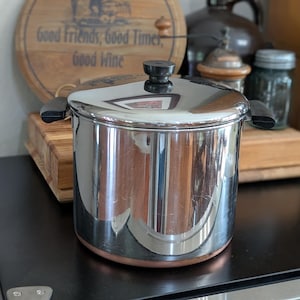May include: A stainless steel pot with a lid and black handles. The pot has a shiny, reflective surface and a copper-colored base. The pot is sitting on a black surface.