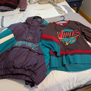 May include: A purple and teal jacket and a brown hockey jersey with the word "Vipers" and a snake logo. The jacket has a hood and the jersey has long sleeves. Both items are on a bed.