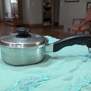 May include: A small, stainless steel saucepan with a black handle and lid. The lid has a black knob. The pan is sitting on a light blue surface.