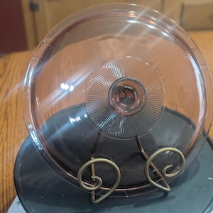 May include: A clear, pink-tinted glass lid with a central knob and radial design. The lid rests on a dark gray, circular base supported by a decorative, gold-colored metal stand. The lid is likely for a casserole dish or similar cookware.