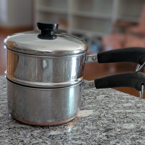 May include: A stainless steel double boiler with a black handle on each side. The lid has a black knob. The pot is sitting on a speckled gray countertop. The background is blurred.