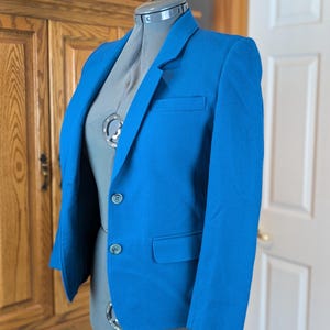 May include: A vibrant blue blazer with a classic two-button closure and a notched lapel. The jacket features a chest pocket and two front pockets. The blazer is displayed on a gray dress form, showcasing its tailored fit and design.