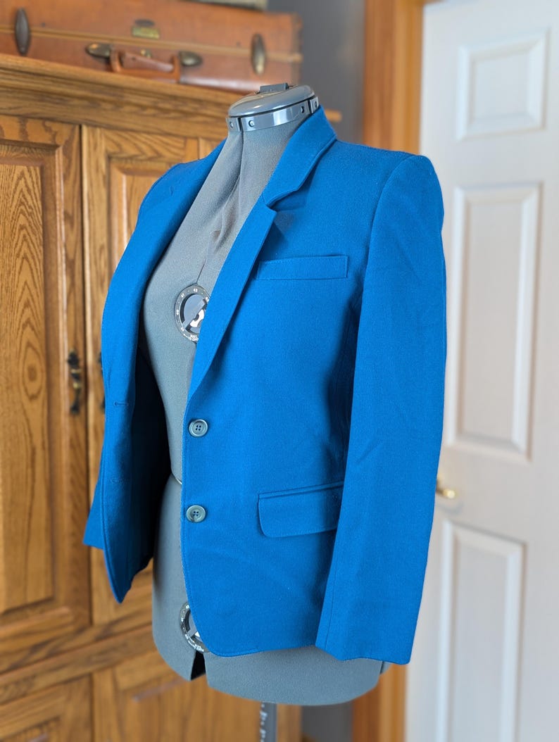 May include: A vibrant blue blazer with a classic two-button closure and a notched lapel. The jacket features a chest pocket and two front pockets. The blazer is displayed on a gray dress form, showcasing its tailored fit and design.