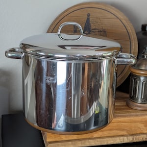 May include: A stainless steel cooking pot with a lid and two side handles. The pot is reflective, with a shiny surface. The lid has a single handle. The pot is sitting on a wooden surface.
