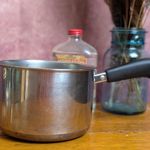 May include: A stainless steel saucepan with a smooth, polished finish and a single black handle. The handle features a small metal loop at the end for hanging. The pot has a cylindrical shape with a flat base and a wide, open top rim.