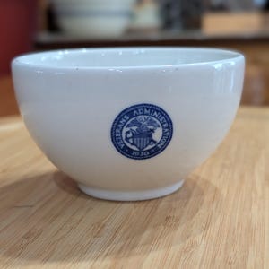 May include: A white ceramic bowl with a rounded shape. A blue emblem with the words "Veterans Administration 1930" is on the side. The bowl is sitting on a wooden surface.