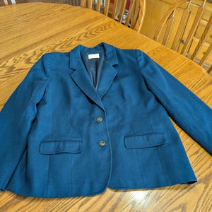 May include: A dark teal blazer with two gold buttons and two front pockets. The blazer has a notched collar and is made of a smooth fabric. The garment is laid flat on a wooden table.