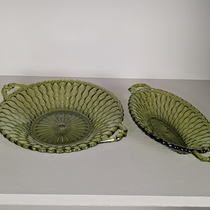 May include: Two olive green glass serving dishes. One is round with handles, the other is oval with handles. Both have a textured, ridged design and a central starburst pattern. The dishes are likely vintage and suitable for serving food.