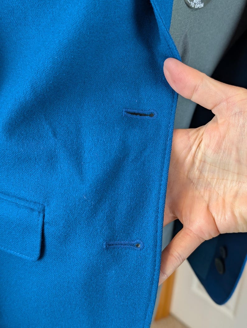 May include: Close-up of a teal blue blazer with a single button closure. The blazer has a notched lapel and a small pocket. The fabric appears to be a smooth, woven material. The blazer is part of a suit.
