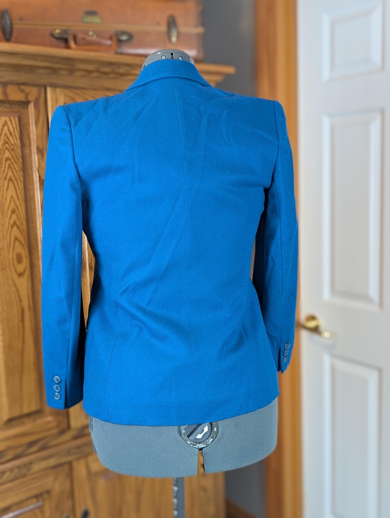 May include: A bright blue blazer displayed on a dress form. The blazer has a classic tailored design with a notched lapel and three buttons on each cuff. The back view shows a fitted silhouette.