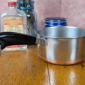 May include: A stainless steel saucepan with a copper-colored base and a black handle. The handle features a silver-colored metal loop at the end. The pan has a cylindrical shape with a smooth, polished finish on the sides and a flat bottom.