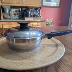 Puede incluir: Una cacerola de acero inoxidable con tapa y asa negra se encuentra sobre una bandeja redonda de madera. La cacerola es reflectante y la tapa tiene un pomo negro. La bandeja es de madera clara, y el fondo incluye un armario de madera.