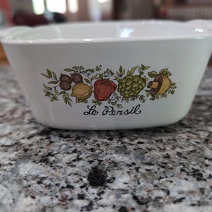 May include: White ceramic bowl with handles, decorated with a colorful illustration of vegetables and the text "Le Persil". The bowl is rectangular with slightly curved sides.