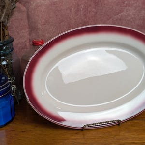 May include: An oval ceramic serving platter with a glossy cream-colored center and a wide, deep burgundy red rim. The platter is displayed on a small metal stand, showcasing its smooth, reflective surface and distinct color contrast.