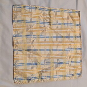 May include: A square pocket square with a plaid pattern in yellow, blue, and white. The fabric has a slight sheen, and the pattern is a classic design. The pocket square is folded and appears to be made of a lightweight material.