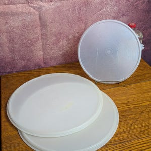 May include: Three round, semi-translucent plastic lids with a smooth, flat surface and a slightly raised outer rim. Two of the lids are stacked horizontally, while the third lid is positioned vertically behind them, supported by a small metal stand.