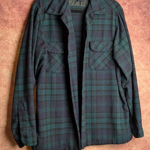 May include: A long-sleeve button-up shirt featuring a dark navy blue and forest green plaid pattern. The shirt includes a pointed collar, two chest pockets with buttoned flaps, and a Pendleton brand label visible on the inner neck lining.