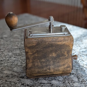 May include: A vintage manual coffee grinder with a square wooden body featuring a dark brown stained finish. The top consists of a silver-toned metal plate with a hinged lid, a small adjustment knob, and a crank handle with a rounded wooden knob.