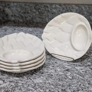 May include: A set of white ceramic appetizer plates with a leaf-like design. Each plate has a central dip bowl. One plate is on a metal stand. The plates are stacked in a neat pile, ready for serving.