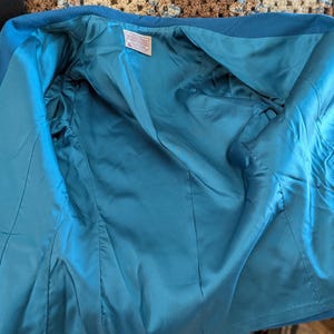 May include: Open view of a teal blue blazer, revealing a matching teal satin lining. The jacket is made of a textured fabric and features a classic tailored design. Tags are visible inside the jacket.