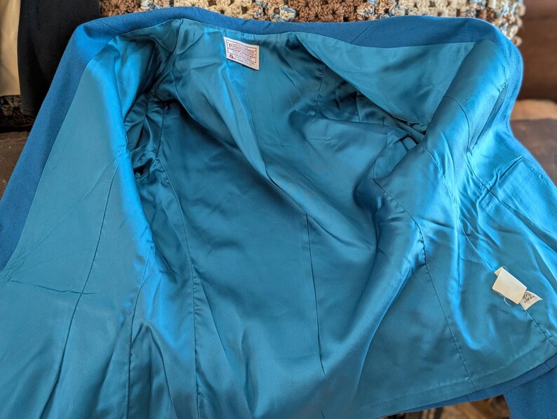 May include: Open view of a teal blue blazer, revealing a matching teal satin lining. The jacket is made of a textured fabric and features a classic tailored design. Tags are visible inside the jacket.