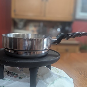May include: A stainless steel saucepan with a black handle rests on a small, black, three-legged stand. The pan is round with a shiny, reflective surface. The stand is placed on a table covered with a floral patterned cloth. The background includes a red wall and wooden cabinets.