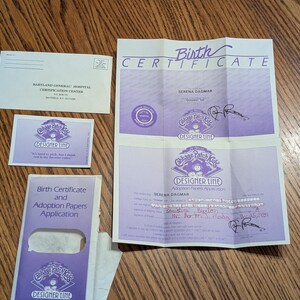 May include: A collection of Cabbage Patch Kids Designer Line paper documents including a white envelope from Babyland General Hospital, a small purple card, a birth certificate and adoption papers application, and a large purple birth certificate.