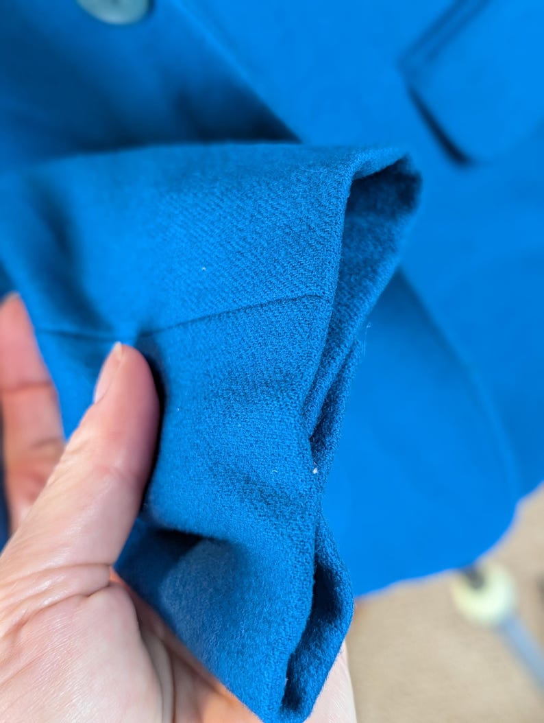 May include: Close-up of a bright blue blazer sleeve. The fabric is a solid color with a textured appearance. The sleeve is folded, revealing the interior lining. A button is visible in the background.