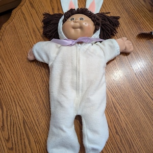 May include: A doll with dark brown yarn hair and a round face wearing a white fleece onesie with an attached hood featuring two white rabbit ears with pink centers. The onesie has a front zipper closure and a light purple ribbon tied at the neck.