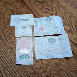May include: A collection of Cabbage Patch Kids paper documents including a Babyland General Hospital Certification Center envelope, a birth certificate, an adoption papers application, and a small card, all featuring pink and green decorative borders.