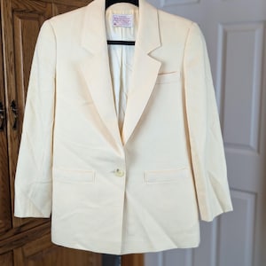 May include: A light yellow blazer with a single-button closure, notched lapels, and two front pockets. The blazer is hanging on a black hanger. The interior lining is a lighter shade of yellow.