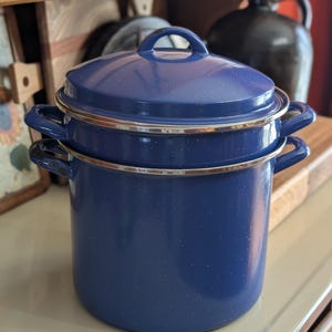 May include: A blue enamelware cooking pot with a lid. The pot has a speckled finish and a silver rim. The lid has a handle. The pot is sitting on a wooden surface. The pot is a classic kitchenware item.
