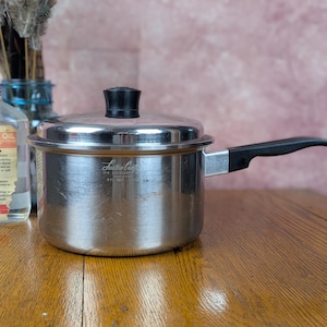May include: A stainless steel saucepan with a matching lid and a black handle. The side of the pot features the engraved text Lustre Craft, The Finest Stainless Steel Cookware, and the pot is positioned next to a clear bottle of mineral oil.