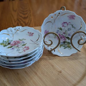 May include: A stack of six white porcelain dessert plates with a floral design, featuring pink and purple roses and green leaves. The plates have a scalloped edge with gold trim. A matching plate is displayed in a gold-colored metal stand.