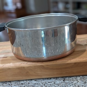 May include: A stainless steel cooking pot with a polished silver finish and a copper-colored base. The pot features two black heat-resistant side handles and a wide, open top with a rolled rim, designed for stovetop use in a kitchen setting.