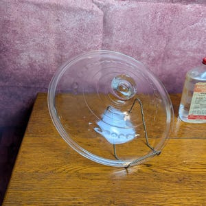 May include: A clear glass circular lid featuring a central knob handle and a decorative embossed pattern of small circles along the outer rim. The lid is supported by a thin metal wire stand and is positioned next to a bottle of heavy mineral oil.