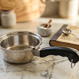 May include: A stainless steel saucepan with a black handle sits on a marble countertop. A vintage cookbook, a small bowl with a wooden spoon, and a measuring spoon are nearby. A small jar is in the background.