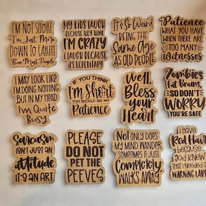 May include: A set of 12 wooden signs with humorous sayings. The signs are all rectangular and have a light brown wood finish. The sayings are printed in black ink and include phrases like "I'm not short, I'm just more down to earth than most people" and "Sarcasm isn't just an attitude, it's an art."