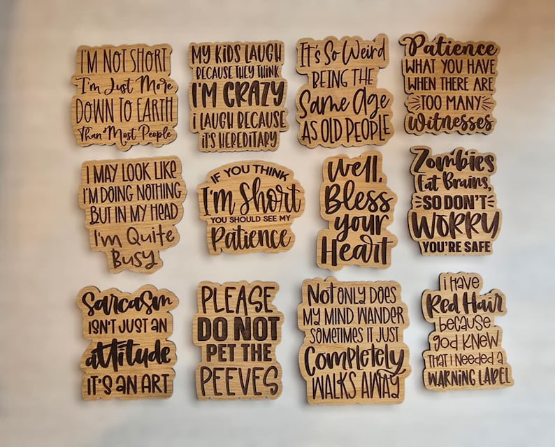 May include: A set of 12 wooden signs with humorous sayings. The signs are all rectangular and have a light brown wood finish. The sayings are printed in black ink and include phrases like "I'm not short, I'm just more down to earth than most people" and "Sarcasm isn't just an attitude, it's an art."