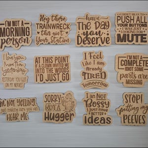 Puede incluir: Un conjunto de doce letreros decorativos de madera con frases humor&iacute;sticas grabadas. Los letreros de color marr&oacute;n claro presentan texto marr&oacute;n oscuro. Las frases incluyen "No soy una persona de la ma&ntilde;ana" y "No soy mandona, solo tengo mejores ideas".