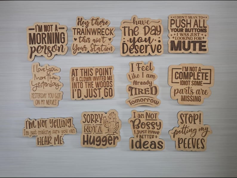 Puede incluir: Un conjunto de doce letreros decorativos de madera con frases humor&iacute;sticas grabadas. Los letreros de color marr&oacute;n claro presentan texto marr&oacute;n oscuro. Las frases incluyen "No soy una persona de la ma&ntilde;ana" y "No soy mandona, solo tengo mejores ideas".
