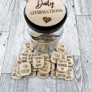 Pode incluir: Um frasco de vidro com uma tampa de madeira que diz "Daily Affirmations". O frasco est&aacute; cheio de pequenos discos de madeira com afirma&ccedil;&otilde;es positivas escritas neles.