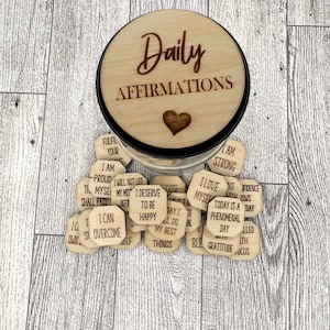Pode incluir: Um pote de madeira com uma tampa que diz "Daily Affirmations" e um cora&ccedil;&atilde;o. O pote est&aacute; cheio de pequenas fichas de madeira com afirma&ccedil;&otilde;es positivas escritas nelas.