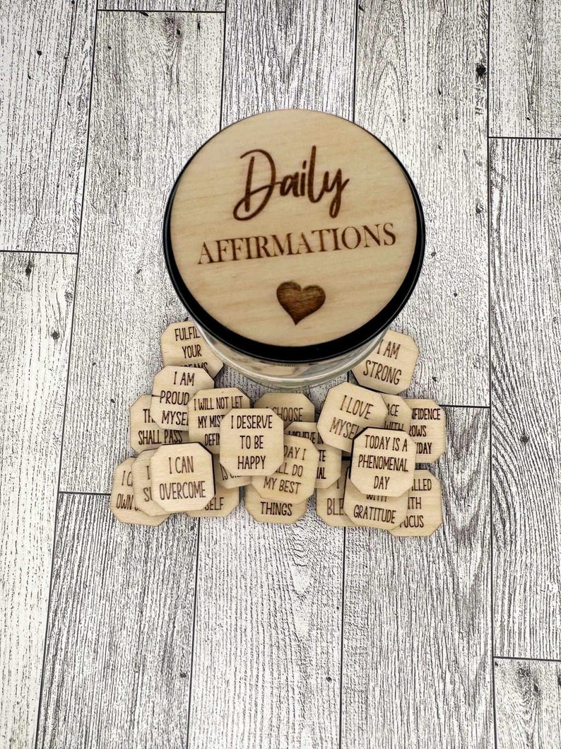 Pode incluir: Um pote de madeira com uma tampa que diz "Daily Affirmations" e um cora&ccedil;&atilde;o. O pote est&aacute; cheio de pequenas fichas de madeira com afirma&ccedil;&otilde;es positivas escritas nelas.