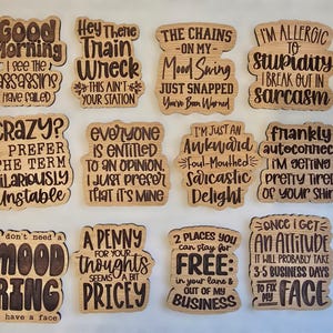 May include: A set of 12 wooden coasters with humorous sayings. The coasters are made of light brown wood and have a laser-etched design. The sayings include "Good Morning I See The Assassins Have Failed", "Hey There Train Wreck This Ain't Your Station", "The Chains On My Mood Swing Just Snapped You've Been Warned", "I'm Allergic To Stupidity I Break Out In Sarcasm", "Crazy? I Prefer The Term Hilariously Unstable", "Everyone Is Entitled To An Opinion I Just Prefer That It's Mine", "I'm Just An Awkward Foul-Mouthed Sarcastic Delight", "Frankly Autoconnect I'm Getting Pretty Tired Of Your Shirt", "I Don't Need A Mood Ring I Have A Face", "A Penny For Your Thoughts Seems A Bit Pricey", "2 Places You Can Stay For Free: In Your Lane & Out Of My Business", "Once I Get An Attitude It Will Probably Take 3-5 Business Days To Fix My Face".