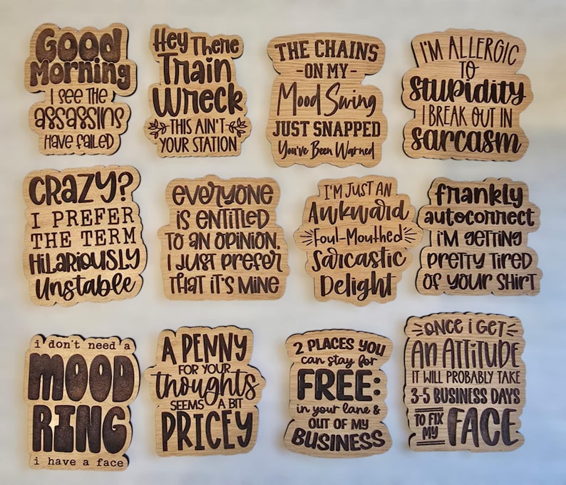 May include: A set of 12 wooden coasters with humorous sayings. The coasters are made of light brown wood and have a laser-etched design. The sayings include "Good Morning I See The Assassins Have Failed", "Hey There Train Wreck This Ain't Your Station", "The Chains On My Mood Swing Just Snapped You've Been Warned", "I'm Allergic To Stupidity I Break Out In Sarcasm", "Crazy? I Prefer The Term Hilariously Unstable", "Everyone Is Entitled To An Opinion I Just Prefer That It's Mine", "I'm Just An Awkward Foul-Mouthed Sarcastic Delight", "Frankly Autoconnect I'm Getting Pretty Tired Of Your Shirt", "I Don't Need A Mood Ring I Have A Face", "A Penny For Your Thoughts Seems A Bit Pricey", "2 Places You Can Stay For Free: In Your Lane & Out Of My Business", "Once I Get An Attitude It Will Probably Take 3-5 Business Days To Fix My Face".