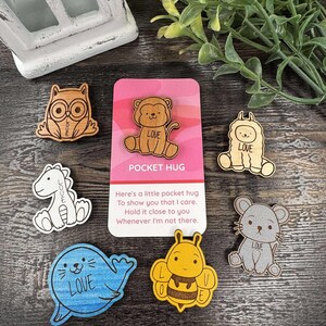 K&ouml;nnte beinhalten: Eine Sammlung von Holz-Taschenumarmungen mit Tierdesigns: Eule, L&ouml;we, Lama, Dinosaurier, Seehund, Biene und Maus. Jedes Tier tr&auml;gt das Wort "LOVE" oder &Auml;hnliches. Eine rosa Karte zeigt "POCKET HUG" und eine Nachricht.