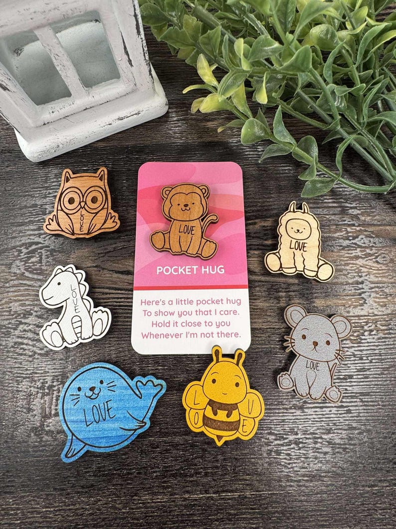 K&ouml;nnte beinhalten: Eine Sammlung von Holz-Taschenumarmungen mit Tierdesigns: Eule, L&ouml;we, Lama, Dinosaurier, Seehund, Biene und Maus. Jedes Tier tr&auml;gt das Wort "LOVE" oder &Auml;hnliches. Eine rosa Karte zeigt "POCKET HUG" und eine Nachricht.