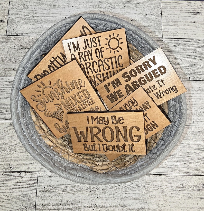 K&ouml;nnte beinhalten: Ein Satz von acht Holzschildern mit humorvollen Spr&uuml;chen. Die Schilder sind aus hellem braunem Holz gefertigt und haben eine abgenutzte Oberfl&auml;che. Die Spr&uuml;che lauten: "Ich bin nur ein Strahl sarcastischen Sonnenscheins", "Es tut mir leid, wir haben uns gestritten", "Sonnenschein gemischt mit einem kleinen Hurrikan", "Ich kann falsch liegen, aber ich bezweifle es", "Dotty", "Heute habe ich es gegessen", "Der Tag, an dem ich weggegangen bin", und "Ugh".