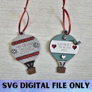Op de afbeelding: Twee houten luchtballonornamenten met rode linten. Eén is grijs met rode harten en sneeuwvlokken, de tekst luidt "May your heart take flight this Christmas!". De andere is blauwgroen met harten en de tekst "Floating into the holiday spirit!"